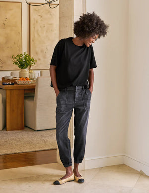 A person stands indoors with hands in the utility pockets of FRANK & EILEEN’s Jameson Jogger Utility Jean in 1981 Wash. They pair it with a black top and tan ballet flats with black toe caps. A dining table and chairs are visible behind them.
