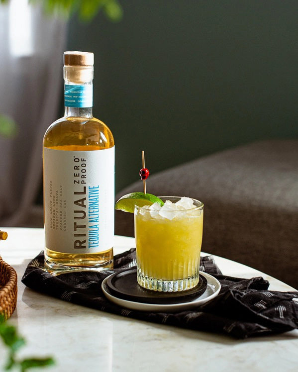 A clear glass bottle of RITUAL ZERO PROOF-TEQUILA ALTERNATIVE by RITUAL ZERO PROOF, a non-alcoholic tequila with a cork top and white label featuring bold black and blue text on a plain background.