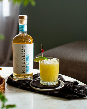 A clear glass bottle of RITUAL ZERO PROOF-TEQUILA ALTERNATIVE by RITUAL ZERO PROOF, a non-alcoholic tequila with a cork top and white label featuring bold black and blue text on a plain background.