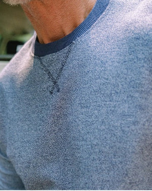 A man with short brown hair wearing the CRIQUET - French Terry Crewneck Sweatshirt in Dusty Blue and white pants stands with one hand in his pocket, looking to his left against a white background.