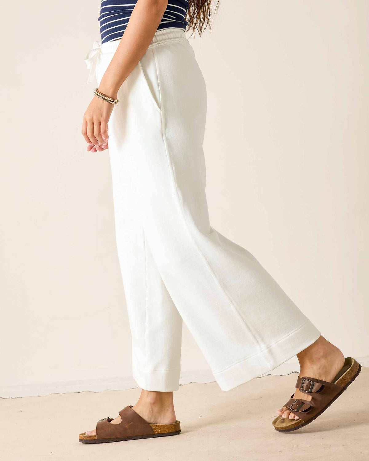 A person stands on a sidewalk in front of urban buildings and a street, wearing a white sweatshirt paired with the MERSEA - FRENCHIE WIDE LEG PANT IN PERIWINKLE from MERSEA. The relaxed fit French terry pants feature a wide-leg silhouette and are styled with sporty beige-and-blue sneakers and a cream tote bag with dark trim.