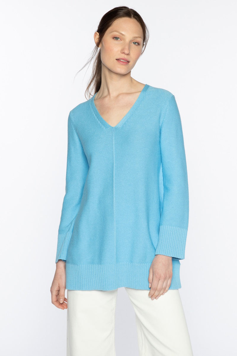 A woman with long hair tied back is wearing the KINROSS CASHMERE - COTTON EASY VEE SWEATER from KINROSS. The light blue V-neck sweater, made of super soft cotton, features ribbed cuffs and hem, and is paired with white pants. She stands against a plain white background, looking slightly towards her right with a neutral and calm expression.