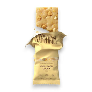 A STARS+HONEY - ICED LEMON COOKIE collagen protein bar in a vibrant yellow wrapper, featuring cookies adorned with white icing and gold sprinkles—some whole, some crumbling—bursting outward against a beige backdrop, capturing a healthy snack from STARS+HONEY packed with delicious flavor.