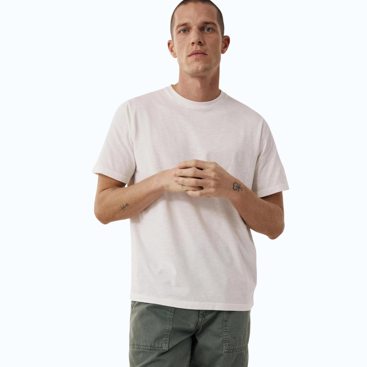 Wearing the HARTFORD - SLUB COTTON CREWNECK T-SHIRT IN OFF WHITE, a short-haired person in green pants stands against a light background with hands loosely clasped, small wrist tattoos visible.
