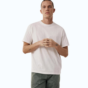 Wearing the HARTFORD - SLUB COTTON CREWNECK T-SHIRT IN OFF WHITE, a short-haired person in green pants stands against a light background with hands loosely clasped, small wrist tattoos visible.