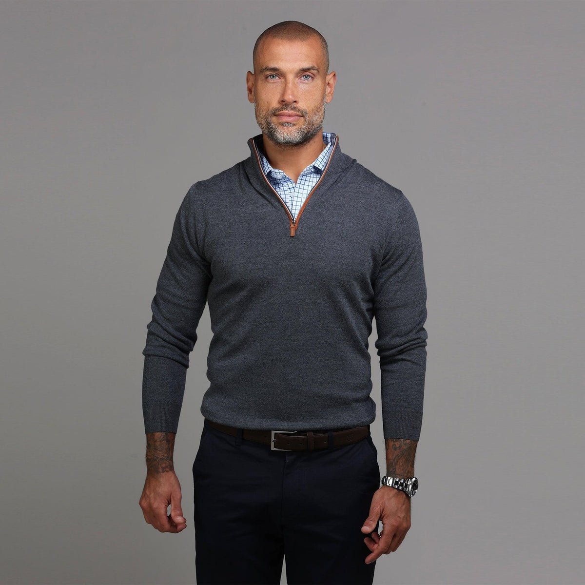 A smiling man with a shaved head and short beard wears the COLLARS AND CO Charcoal with Brown Suede Trim Cashmere Touch Australian Merino Quarter Zip Sweater over a blue checkered shirt, standing against a plain gray background.