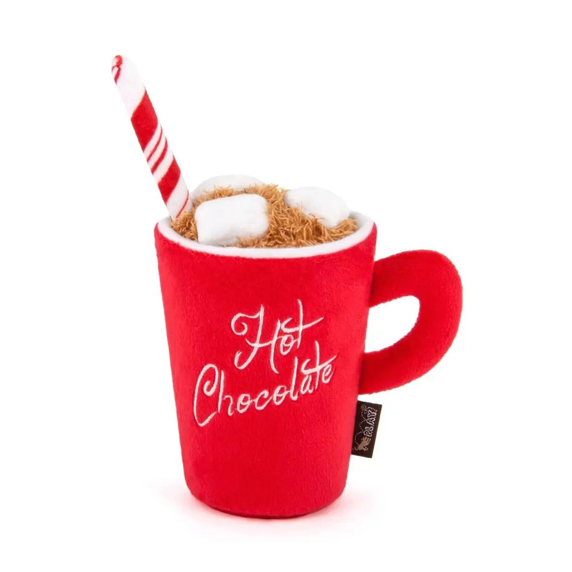 A yellow labrador retriever lies on the floor, drinking from a red HOLIDAY CLASSIC - HOT CHOCOLATE cup toy by PLAY PET LIFESTYLES/BRANDS, complete with a candy cane detail. Beside the pup is a festive holiday dog toy. The cozy indoor scene features a table and chairs, with a Christmas tree glowing in the background.