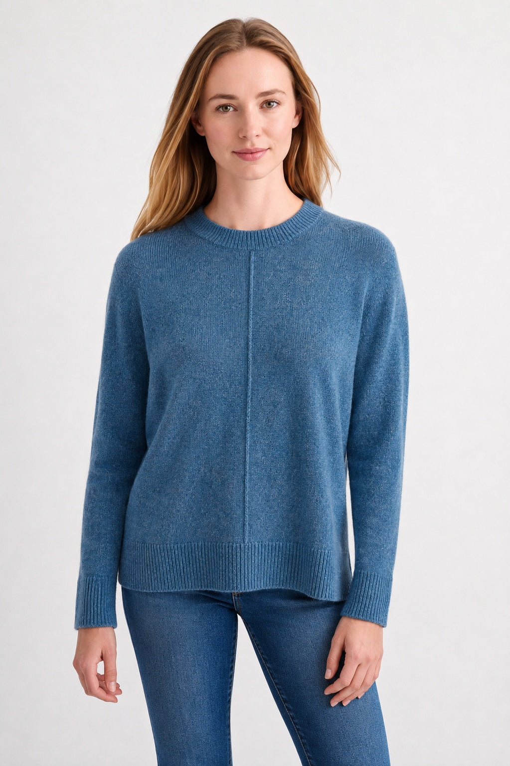 A woman with long, light brown hair wears the Kinross Cashmere Hi Low Seamed Crew Neck Cashmere Sweater in Loch, styled in a relaxed classic fit with blue jeans. She stands against a plain light background, smiling slightly at the camera.