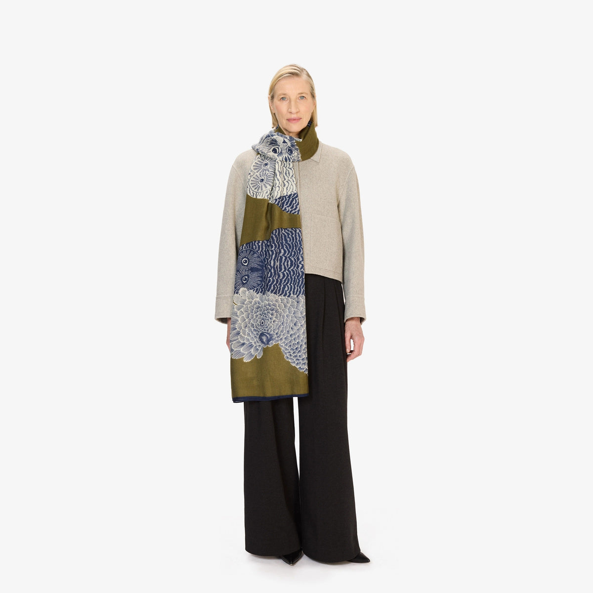 A woman stands against a white background wearing a light gray sweater, black pants, and the INOUI EDITIONS &quot;Owls on a Branch&quot; wool scarf in khaki by INOUI, featuring a blue and white owl design.