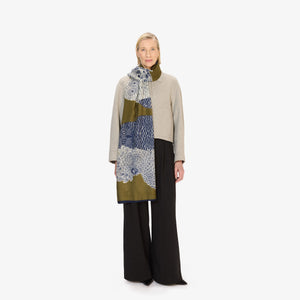 A woman stands against a white background wearing a light gray sweater, black pants, and the INOUI EDITIONS "Owls on a Branch" wool scarf in khaki by INOUI, featuring a blue and white owl design.