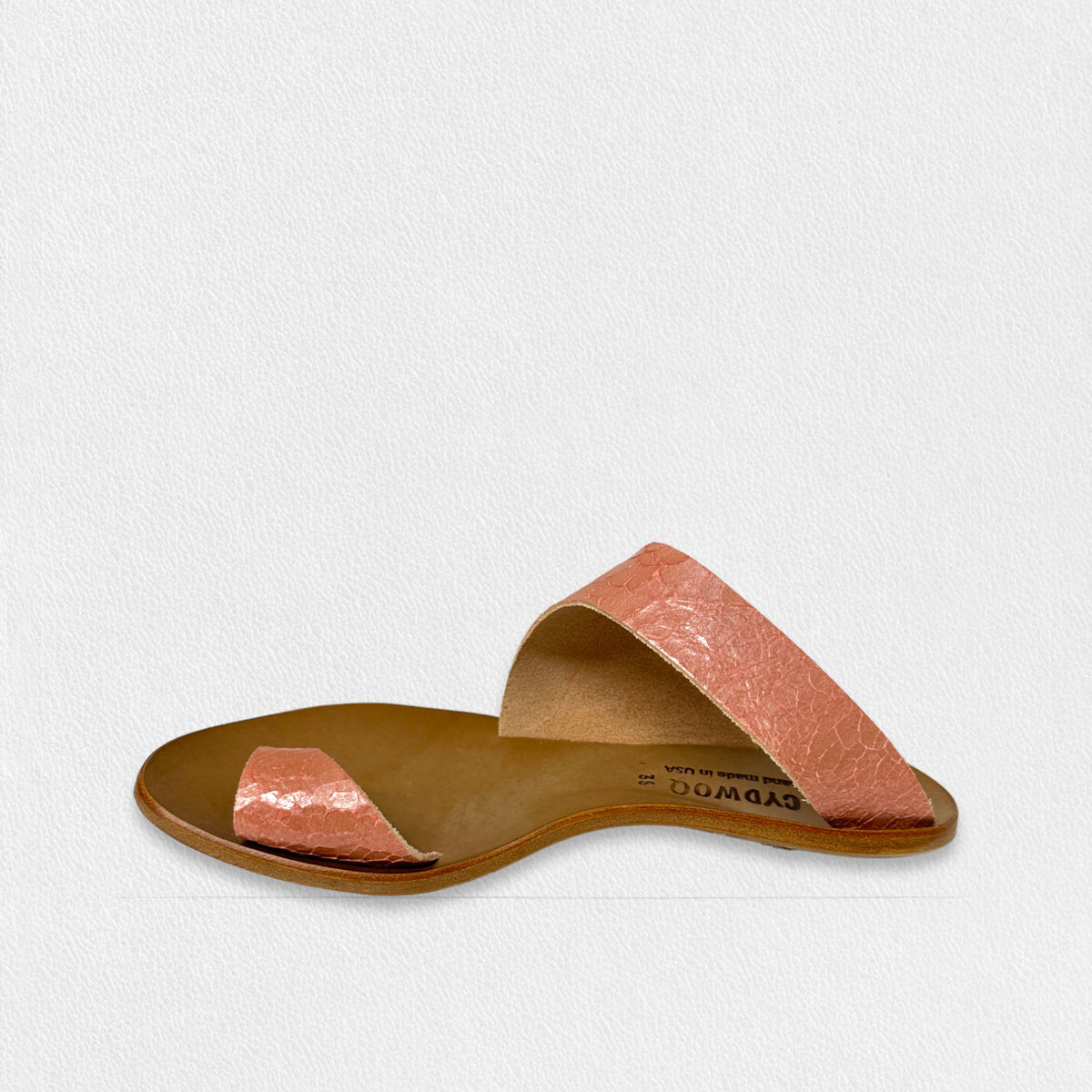 The CYDWOQ SANDAL by CYDWOQ (SIDEWALK) features a single flat slide design with a brown sole and two wide, textured pink straps—one across the toes and another near the instep—highlighting its modern aesthetic against a light background.