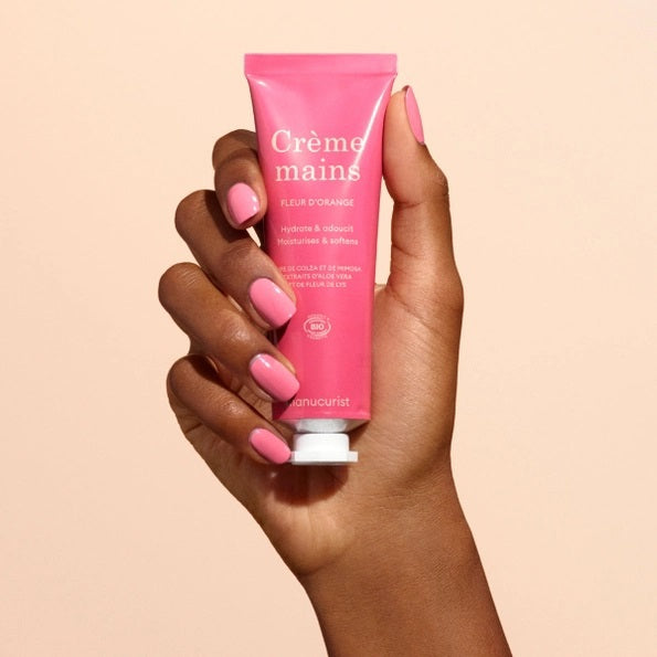 Close-up of two hands with light skin; one is gently applying MANUCURIST - COSMOS ORGANIC HAND CREAM, ORANGE BLOSSOM by MANUCURIST INC to the back of the other against a neutral beige background. Ideal for hand and nail care, this organic moisturizer is formulated with natural ingredients to leave fingers nourished. The nails are perfectly groomed and finished with a subtle pale nude polish for an elegant touch.