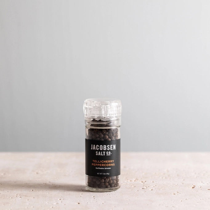 A glass JACOBSEN SALT - GRINDER TELLICHERRY PEPPERCORNS jar from JACOBSEN SALT CO. rests on a light stone surface with a plain gray background, highlighting this premium pre-filled grinder.