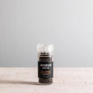 A glass JACOBSEN SALT - GRINDER TELLICHERRY PEPPERCORNS jar from JACOBSEN SALT CO. rests on a light stone surface with a plain gray background, highlighting this premium pre-filled grinder.