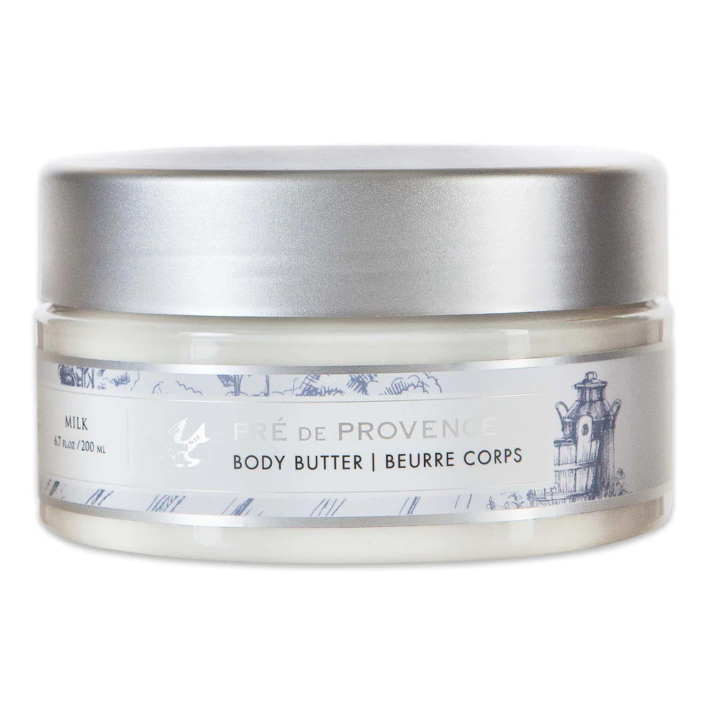The BODY BUTTER - MILK from EUROPEAN SOAPS, LLC is housed in a translucent jar with a silver lid. The body butter is enriched with Shea Butter and features intricate illustrations of a landscape and lighthouse on its label. The label reads &quot;Pré de Provence Body Butter | Beurre Corps&quot; and &quot;Milk.&quot; Perfect for dry skin, the jar contains 6.7 fl oz (200 ml) of this moisturizing product.