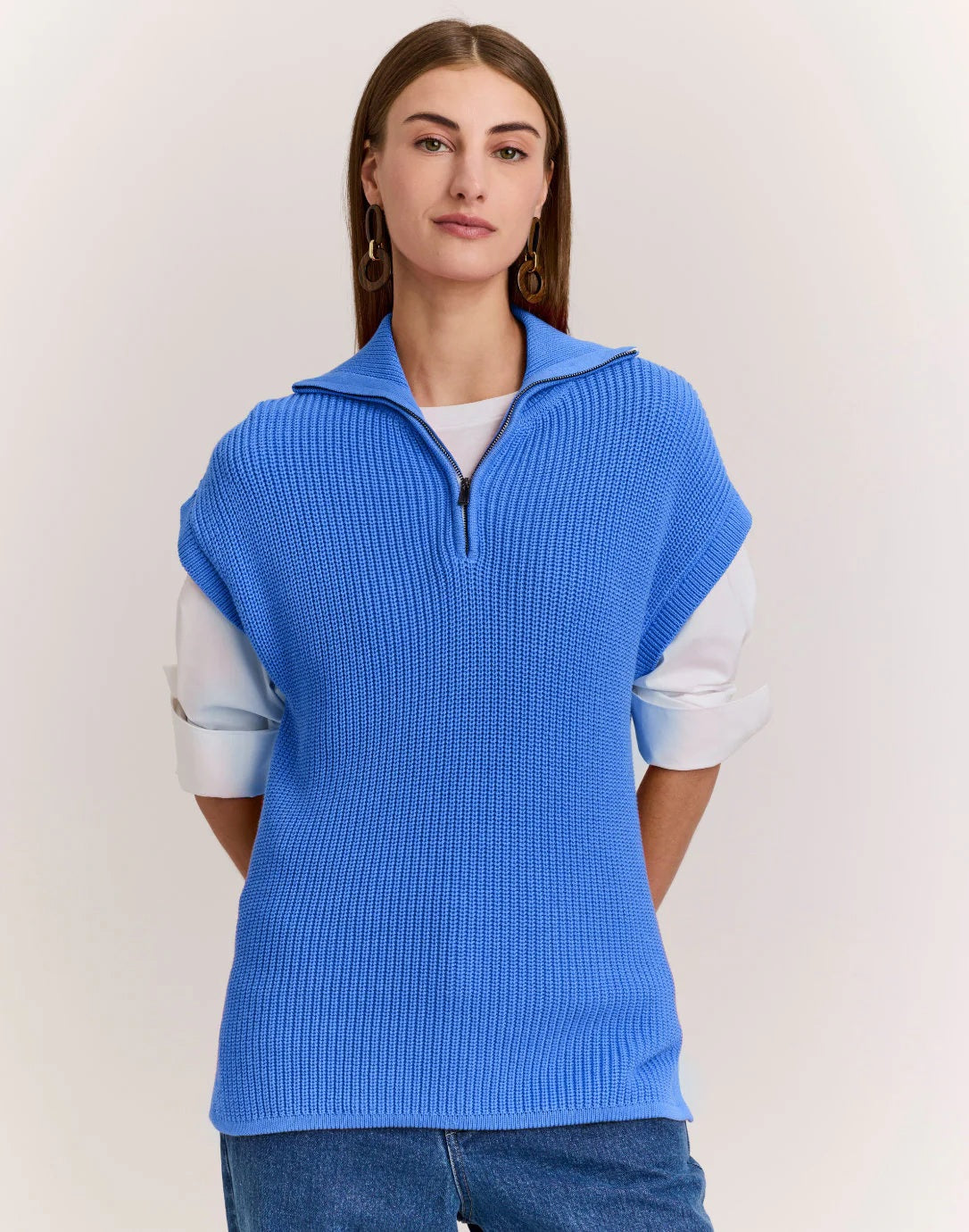 A woman with straight brown hair wears the HINSON WU - BROOKE SLEEVELESS KNIT VEST, a bright blue textured cotton-silk sweater, over a white shirt and blue jeans, standing against a plain light background.
