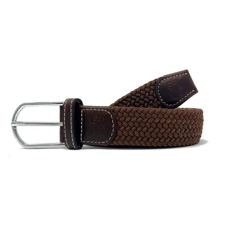 A person wears a black shirt tucked into white pants with hands in pockets, accessorized with the ROOSTAS - MOJAVE WOVEN STRETCH BELT by ROOSTAS and a smartwatch on their left wrist.