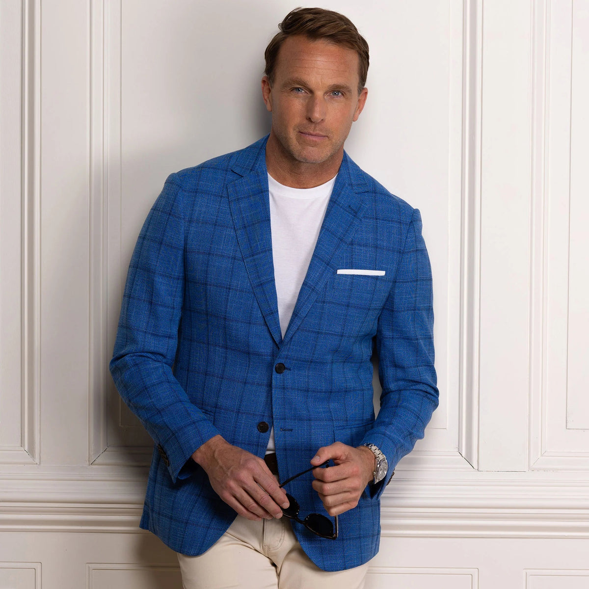 A man wearing sunglasses, the COLLARS AND CO MAVERICK TRAVEL BLAZER IN BLUE WINDOWPANE over a white t-shirt, along with light pants and a brown belt, stands smiling with one hand in his pocket against a white paneled wall.
