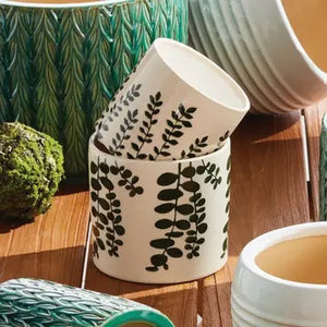 Two NAPA HOME AND GARDEN Maren Pots in white ceramic with an embossed leaf pattern sit on a wooden table, one with a leafy plant. Nearby are two stacked books and a partially visible empty green cachepot.