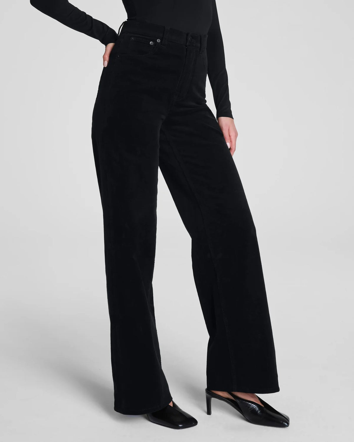 A woman poses against a plain background wearing the SPANX - CORDUROY WIDE LEG JEAN IN BLACK, paired with a fitted black long-sleeve top, black belt, and black heels, her hands in her pockets and expression neutral.