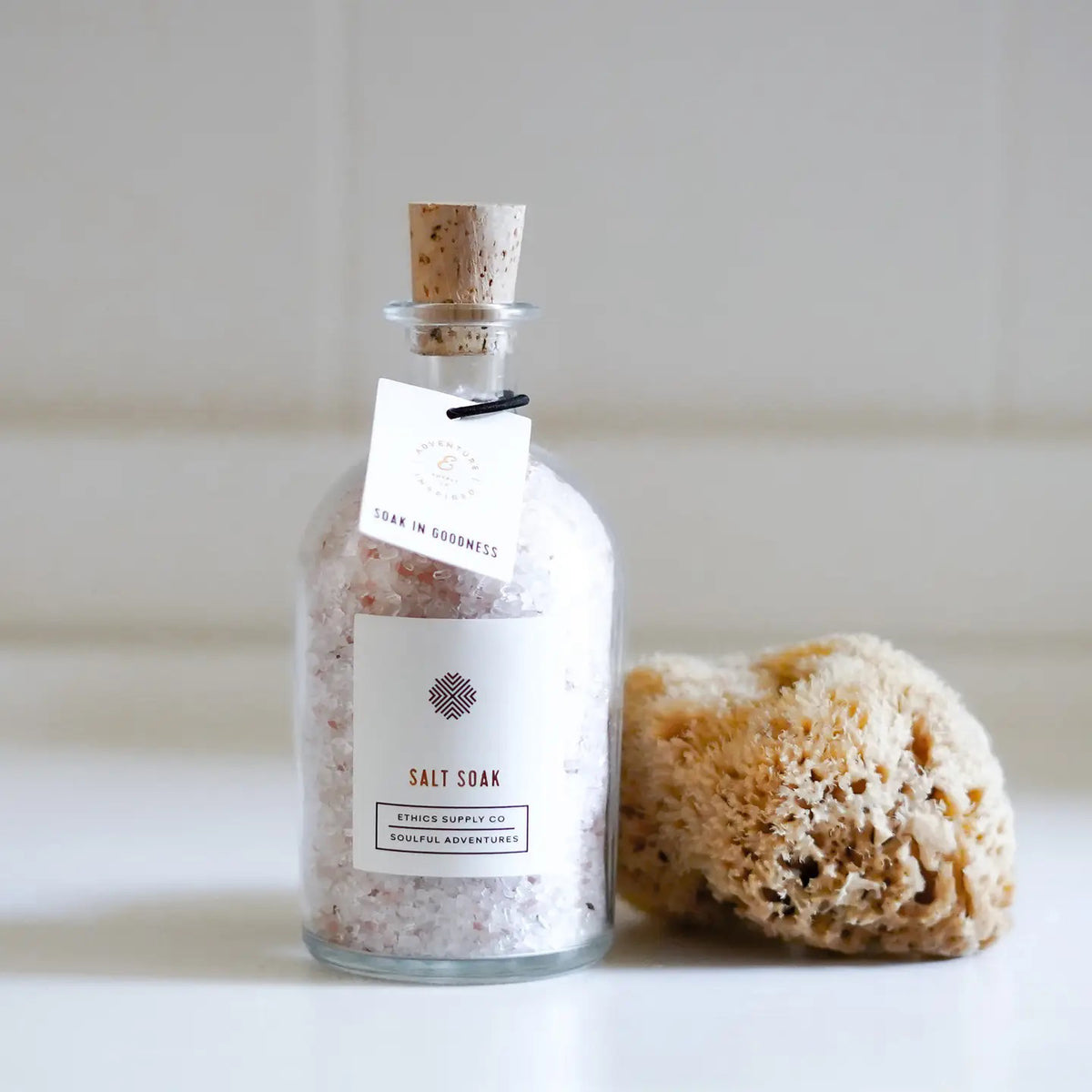 A bottle of ETHICS SUPPLY CO's "Soak in Goodness" Bubbly Bath Salt Soak rests on a wooden table surrounded by lavender and wild violets, while sunlight streams through the window, creating a serene atmosphere.