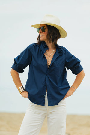 A person stands outdoors with hands on hips, smiling, wearing sunglasses, a wide-brimmed hat, gold jewelry, white pants, and the KMJ SOUTHERN LOVER SHIRT IN NAVY by KMJ. The background is light and neutral.