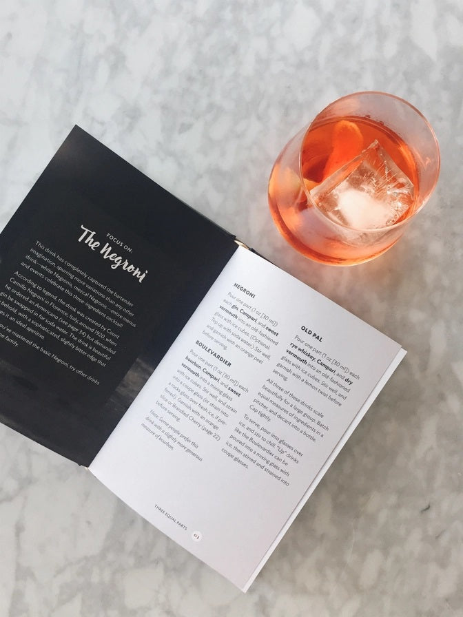 The SHAKE. STIR. SIP. BY KARA NEWMAN recipe book from CHRONICLE BOOKS rests on a light marble surface alongside an orange cocktail with ice, a metal jigger, and a cocktail strainer.
