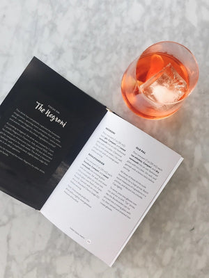 The SHAKE. STIR. SIP. BY KARA NEWMAN recipe book from CHRONICLE BOOKS rests on a light marble surface alongside an orange cocktail with ice, a metal jigger, and a cocktail strainer.