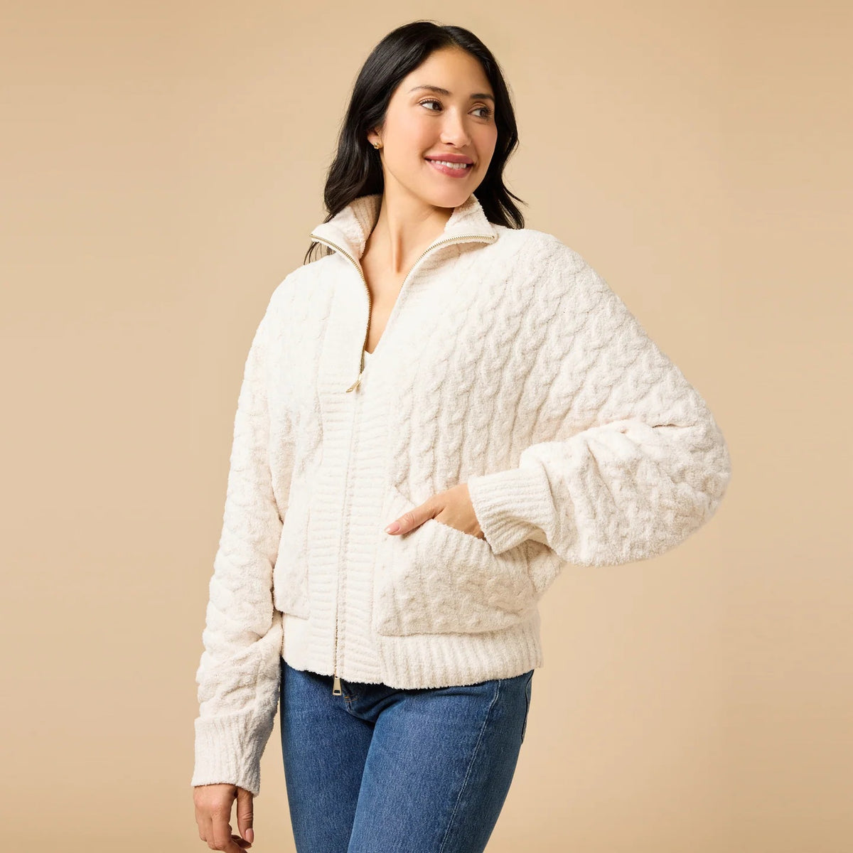 A woman wearing a cream-colored, plush SOFTIES - CABLE KNIT MARSHMALLOW FUNNEL NECK DOUBLE ZIP JACKET with a funnel neck and double zipper, paired with blue jeans.