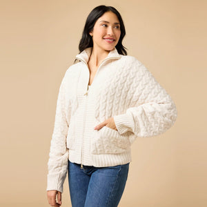 A woman wearing a cream-colored, plush SOFTIES - CABLE KNIT MARSHMALLOW FUNNEL NECK DOUBLE ZIP JACKET with a funnel neck and double zipper, paired with blue jeans.