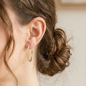 The ELLA STEIN - A HOOP THING EARRINGS by ELLA STEIN are gold-plated hoop earrings adorned with small clear stones along the outer edge, showcased on a plain white background.
