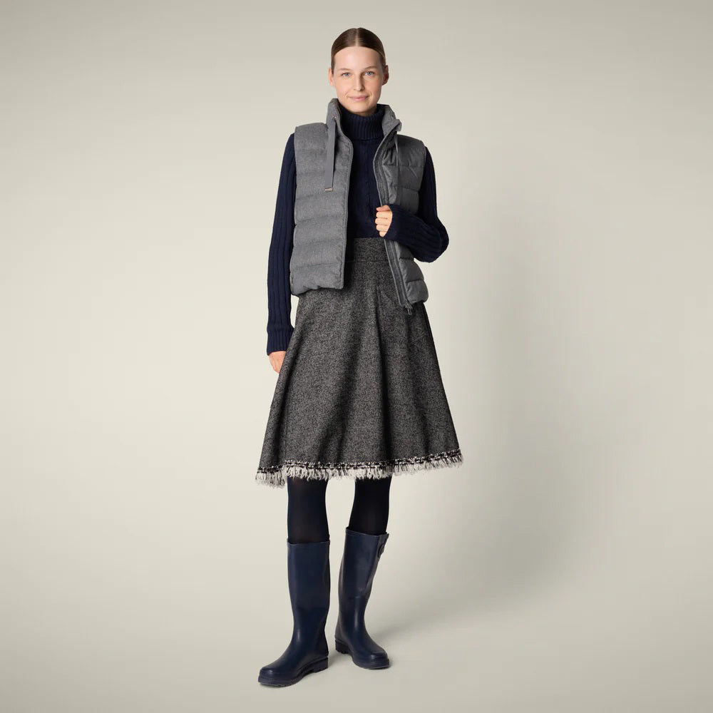 A woman with her hair tied back smiles against a plain background, wearing the SAVE THE DUCK - NYLA PUFFER VEST by SAVE THE DUCK over a navy turtleneck, gray fringed A-line skirt, navy tights, and blue rain boots.