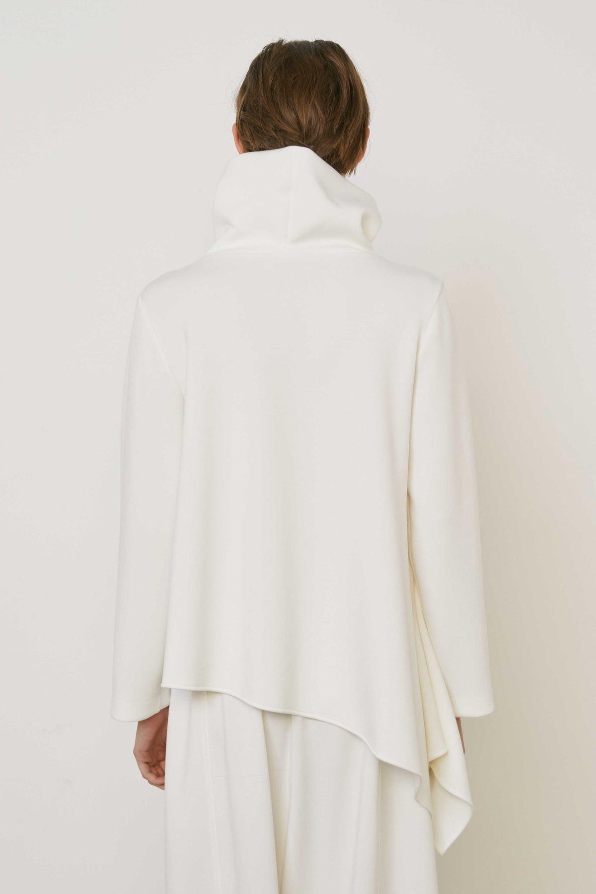 A person with brown hair faces away wearing the MEIMEIJ ASYMMETRICAL COWLNECK TOP in lightweight scuba fabric, featuring an asymmetrical hem, paired with matching white bottoms, against a plain white background.