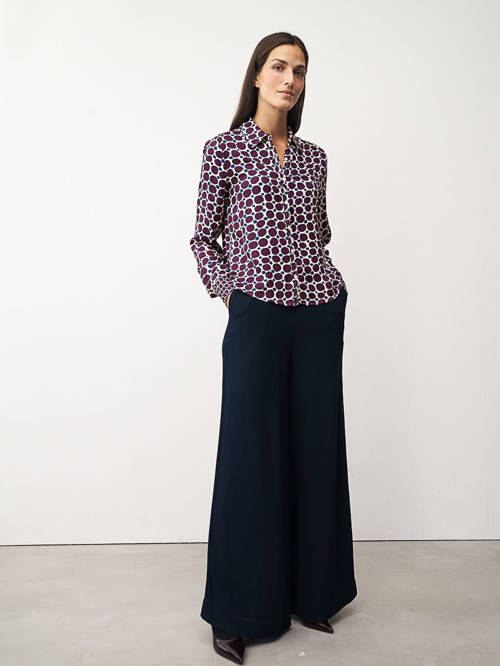 A woman with long brown hair smiles gently, arms crossed, wearing the 0039 ITALY Sia Blouse—a green and white geometric print blouse with pink dots—paired with brown pants against a plain white background.