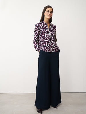 A woman with long brown hair smiles gently, arms crossed, wearing the 0039 ITALY Sia Blouse—a green and white geometric print blouse with pink dots—paired with brown pants against a plain white background.