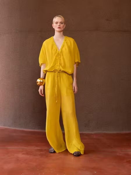 A person with light blonde hair in a low ponytail stands with their back to the camera, wearing the NUDE Tie Waist V Neck Blouse by NUDE in yellow, set against a plain brown background.