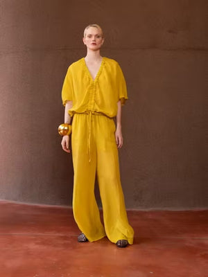 A person with light blonde hair in a low ponytail stands with their back to the camera, wearing the NUDE Tie Waist V Neck Blouse by NUDE in yellow, set against a plain brown background.