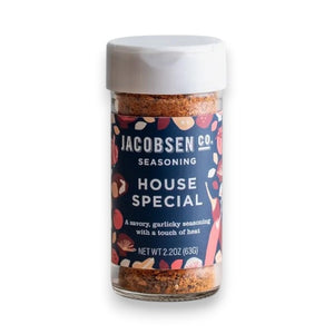 A jar of JACOBSEN SALT CO. HOUSE SPECIAL SEASONING sits on a kitchen counter, surrounded by a ceramic bowl, a glass jar of honey, a bottle of oil, and wooden utensils.