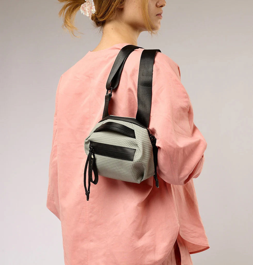 The DANIELLA LEHAVI BONNIE MINI TRAVEL CROSSBODY, a small brown vegan bag with black accents and strap, is displayed on a white pedestal against a plain beige background.