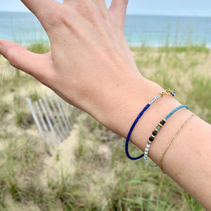 The ALICE RISE - PEARL BRACELET by ALICE RISE features delicate blue, white, green, cream, and gold beads in unique patterns with a 14k gold-filled clasp, shown on a textured blue surface.