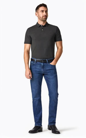 A man stands facing forward, wearing a dark short-sleeve polo, 34 HERITAGE - COURAGE STRAIGHT LEG JEAN IN MID URBAN WASH, a black belt, and black shoes. One hand is in his pocket; he has short hair, a trimmed beard, and a neutral expression.
