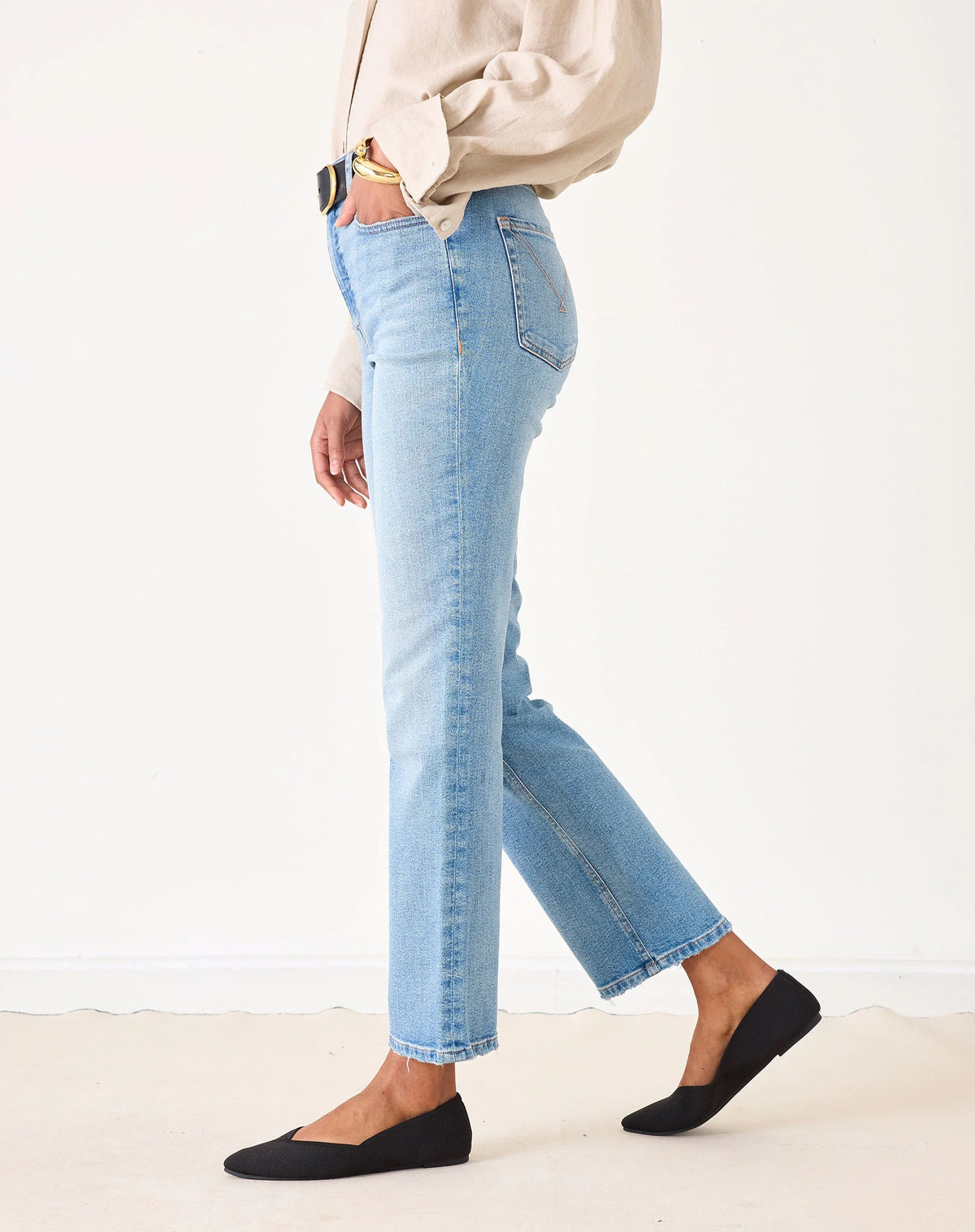 A person wearing a beige button-up shirt, MERSEA Bessie Straight Leg Jean in Caribbean made from recycled cotton, and black flats stands holding a woven straw tote bag with black handles. Their left hand is in their pocket, face not visible.