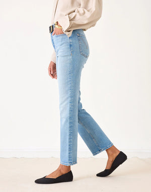 A person wearing a beige button-up shirt, MERSEA Bessie Straight Leg Jean in Caribbean made from recycled cotton, and black flats stands holding a woven straw tote bag with black handles. Their left hand is in their pocket, face not visible.