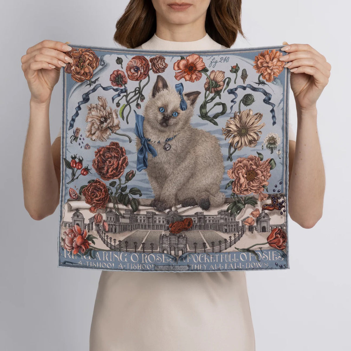 A woman holds the SABINA SAVAGE "Ring a Ring o' Roses" silk twill scarf in Glacier/Ballet, featuring a blue-eyed cat with a ribbon amidst Victorian botanicals and a historic building backdrop, with "Ring a Ring o' Roses, A Pocket Full of Posies" elegantly displayed.