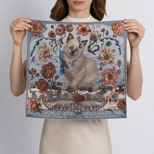 A woman holds the SABINA SAVAGE "Ring a Ring o' Roses" silk twill scarf in Glacier/Ballet, featuring a blue-eyed cat with a ribbon amidst Victorian botanicals and a historic building backdrop, with "Ring a Ring o' Roses, A Pocket Full of Posies" elegantly displayed.