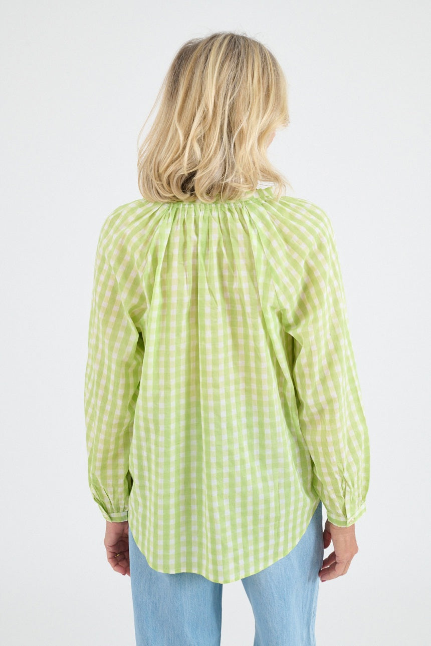 A smiling older woman with blonde hair exudes casual elegance in the STORIATIPIC - IRIS GINGHAM BLOUSE IN GREEN, standing with her hands in her pockets against a plain white background.