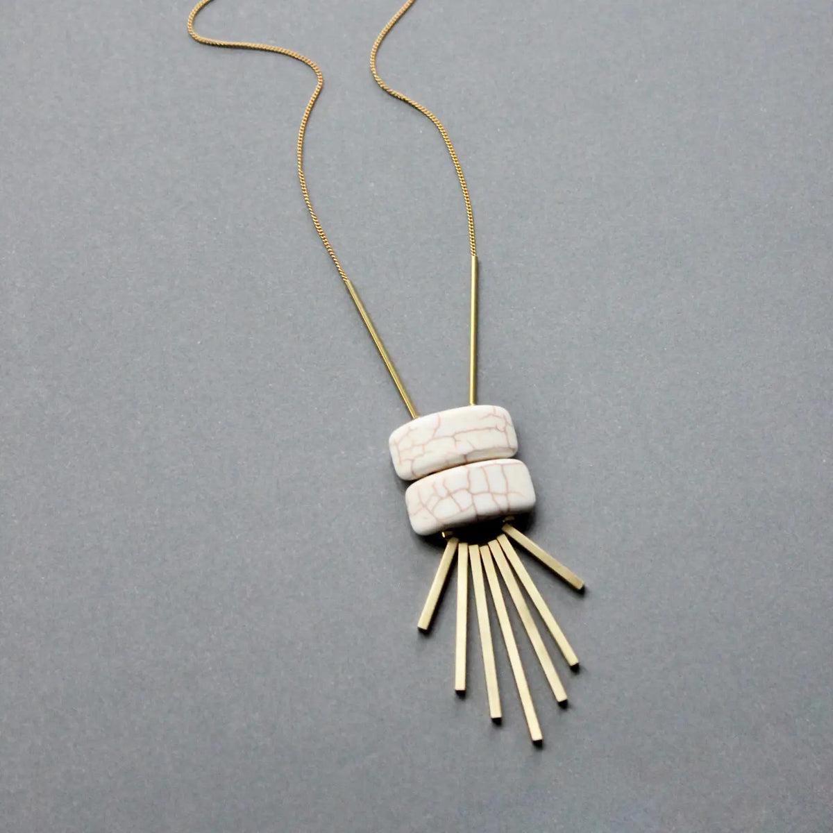 The DAVID AUBREY - WHITE GEOMETRIC PENDANT NECKLACE by DAVID AUBREY showcases two white magnesite beads above six gold-plated brass bars arranged like rays, all set against a simple gray background.