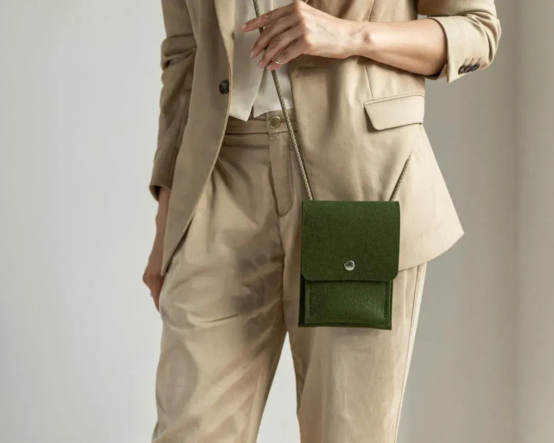A person in a beige sweater and tan pants holds the WOOLBERRY WOOL FELT PHONE BAG, a small gray handcrafted bag with a flap and button closure. The image is cropped at the shoulders and knees.