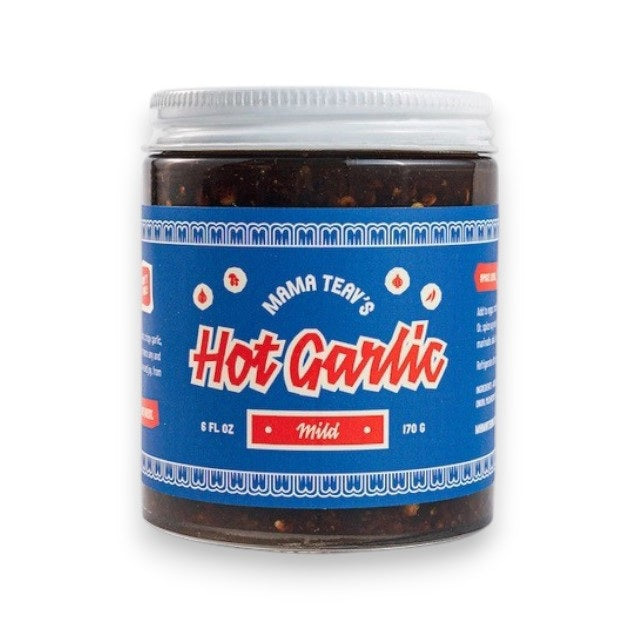 A glass jar of MAMA TEAV’S - MILD HOT GARLIC CHILI CRISPS, featuring a blue and red label, rests on a light gray background. Inside the jar is a dark, chunky condiment from MAMA TEAV’S that’s ideal for anyone looking for a vegan and gluten-free hot garlic chili crisp.
