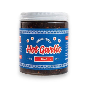 A glass jar of MAMA TEAV’S - MILD HOT GARLIC CHILI CRISPS, featuring a blue and red label, rests on a light gray background. Inside the jar is a dark, chunky condiment from MAMA TEAV’S that’s ideal for anyone looking for a vegan and gluten-free hot garlic chili crisp.
